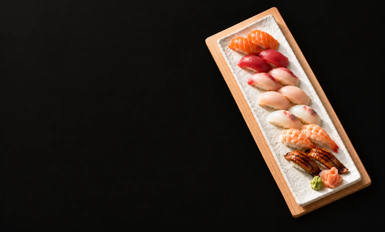 Fourteen pieces of nigiri in seven pairs on a white stone plate on a hinoki board, in soft light on a dark surface.