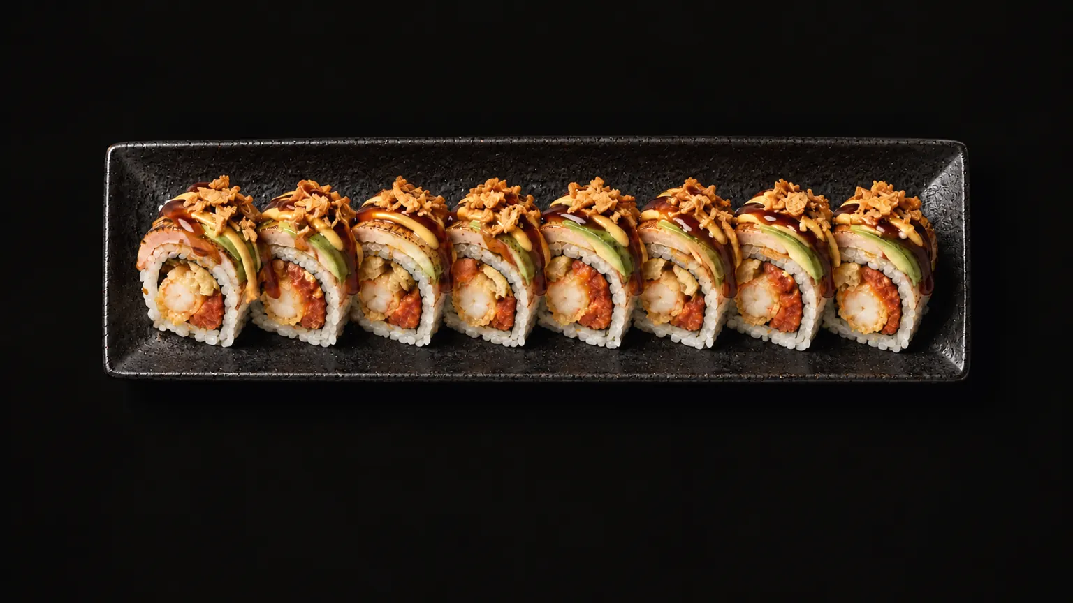 Eight pieces of the Densetsu Roll on a long black plate in a pool of warm light.